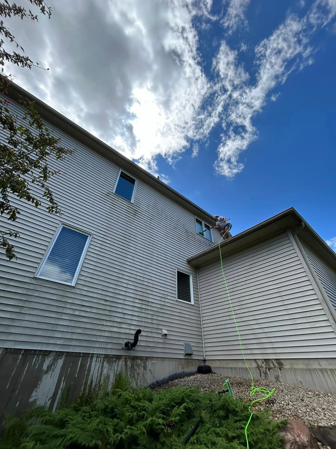 Worker using rope to clean windows on a two-story house under a partly cloudy sky.