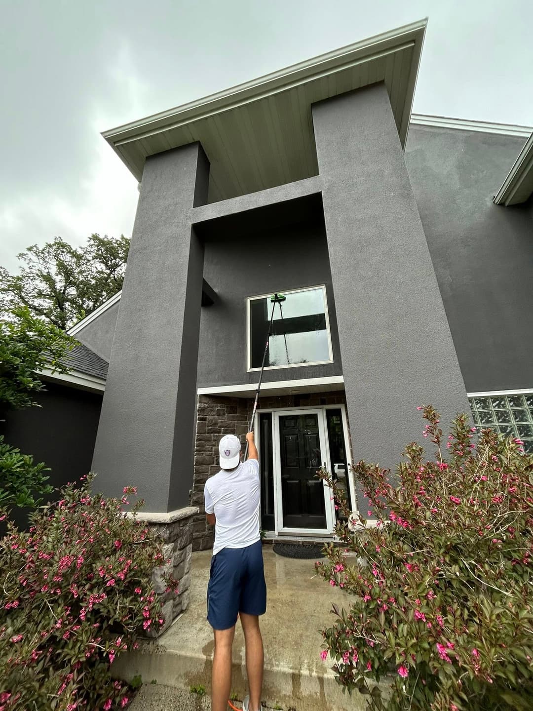 Man cleaning high window of modern gray house with extendable pole, surrounded by greenery.